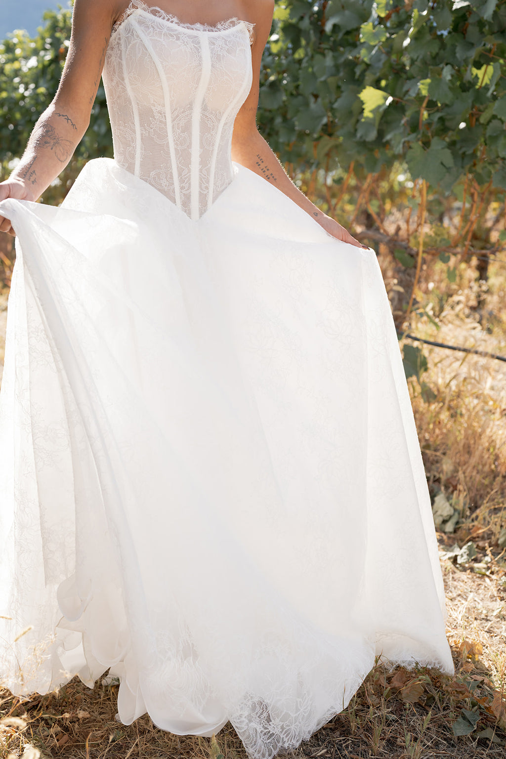 Amanti Contessa white strapless dress with a flowing full A-line skirt in floral Chantilly lace skirt held by a woman in an outdoor setting. Le Reve Boutique, St. Albert Alberta