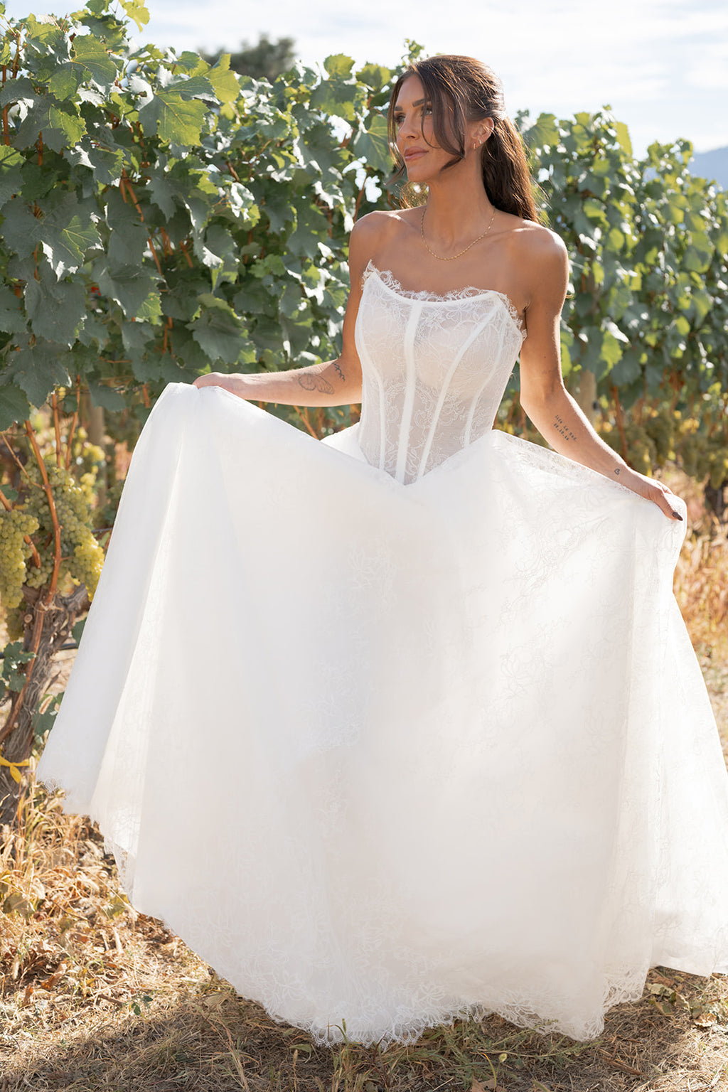 Woman in a white Amanti Contessa wedding dress, Crafted in floral Chantilly lace, this gown blends delicate texture with lightweight movement, making it a perfect choice for brides who want both sophistication and comfort. The zipper back closure ensures a secure fit while maintaining clean lines and a polished finish, standing in a vineyard. Le Reve Boutique, St. Albert Alberta