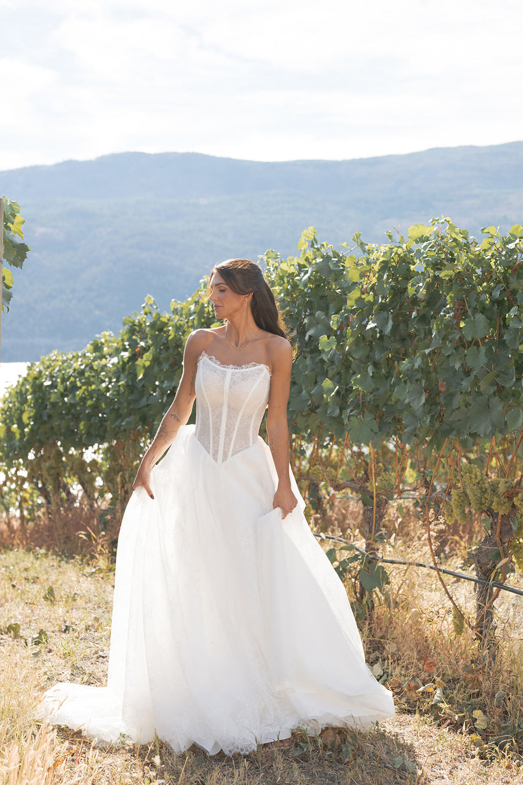 Woman in a white Amanti Contessa wedding dress, with Basque Waist Lace Strapless Wedding Dress with Cat-Eye Neckline,standing in a vineyard with mountains in the background.  Le Reve Boutique, St. Albert Alberta