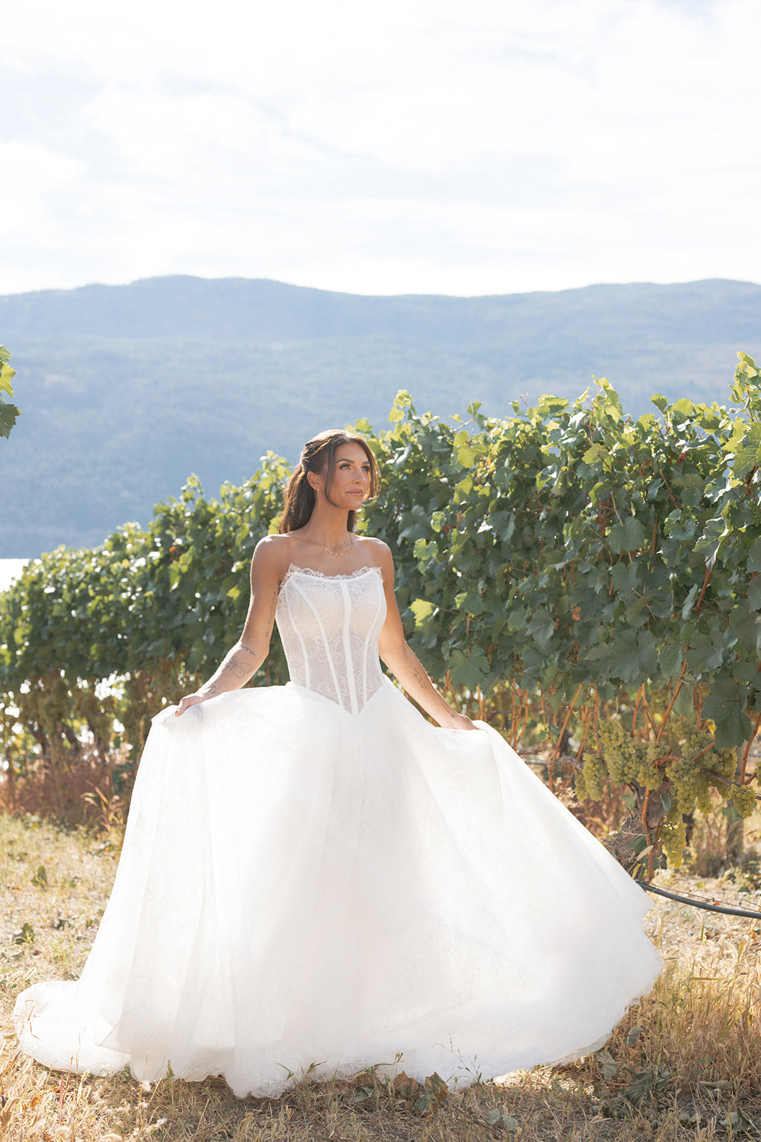 Woman in an Amanti Contessa A-line wedding dress combines a strapless cat-eye neckline and an unlined exposed-boning corset, the bodice creates a striking silhouette while offering a touch of daring elegance. The basque waist elongates the torso and flatters the figure, flowing seamlessly into a full A-line skirt white wedding dress standing in a vineyard with mountains in the background.  Le Reve Boutique. Edmonton, Alberta