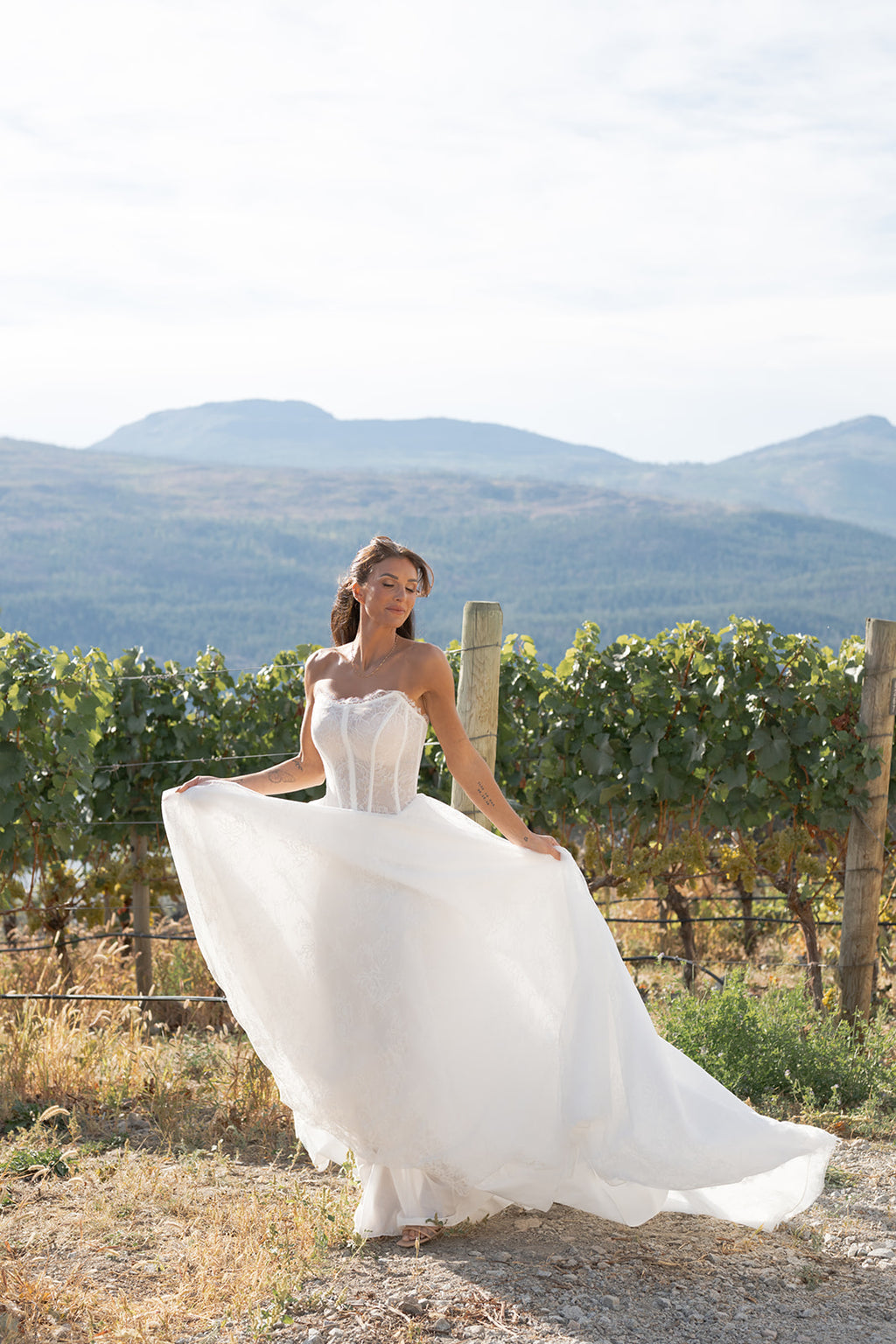 Woman in an Amanti Contessa white wedding dress standing in a vineyard with mountains in the background.  This A-line wedding dress combines a strapless cat-eye neckline and an unlined exposed-boning corset, the bodice creates a striking silhouette while offering a touch of daring elegance. The basque waist elongates the torso and flatters the figure, flowing seamlessly into a full A-line skirt.  Le Reve Boutique. St. Albert, Alberta
