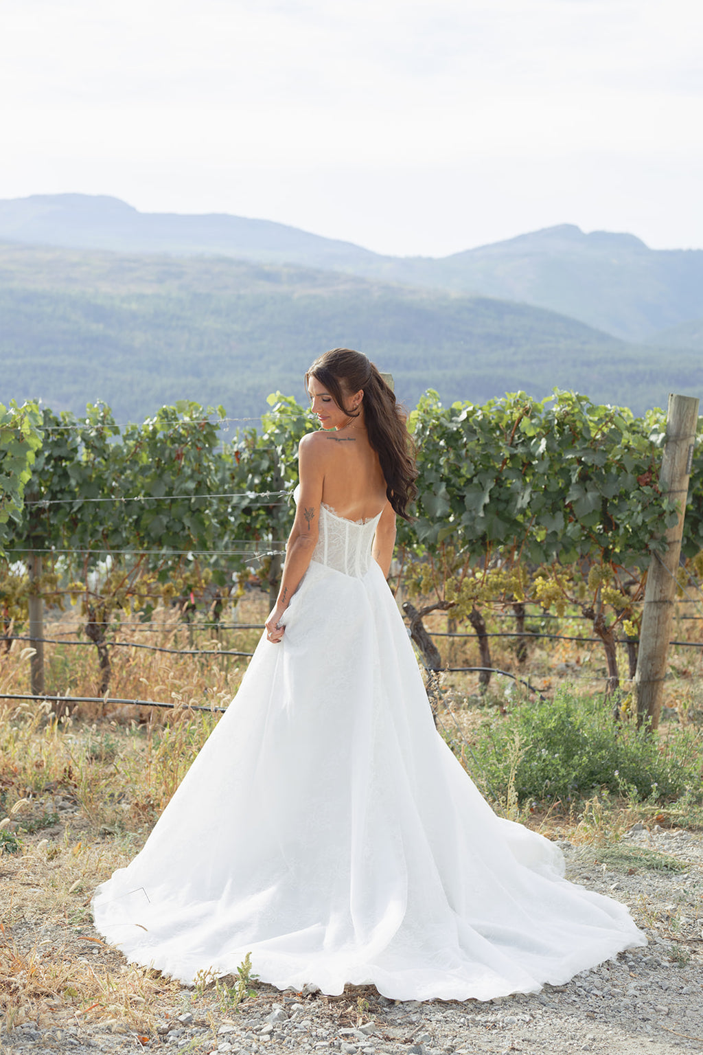 Woman in a white Full A-line skirt Amanti Contessa wedding dress in floral Chantilly lace. Zipper back closure for ease and elegance wedding dress standing in a vineyard with mountains in the background.  Le Reve Boutique. St. Albert, Alberta