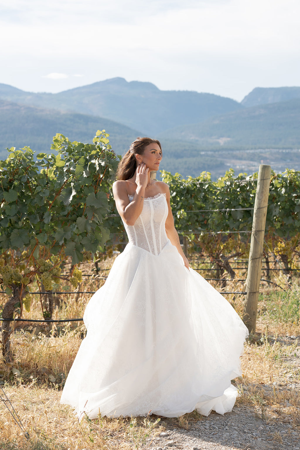 Woman in a white Amanti Contessa Strapless cat-eye neckline wedding dress with exposed boning corset. Basque waistline for a flattering, elongated shape. Full A-line skirt in floral Chantilly lace.  Zipper back closure for ease and elegance, standing in a vineyard with mountains in the background. Le Reve Boutique, Edmonton