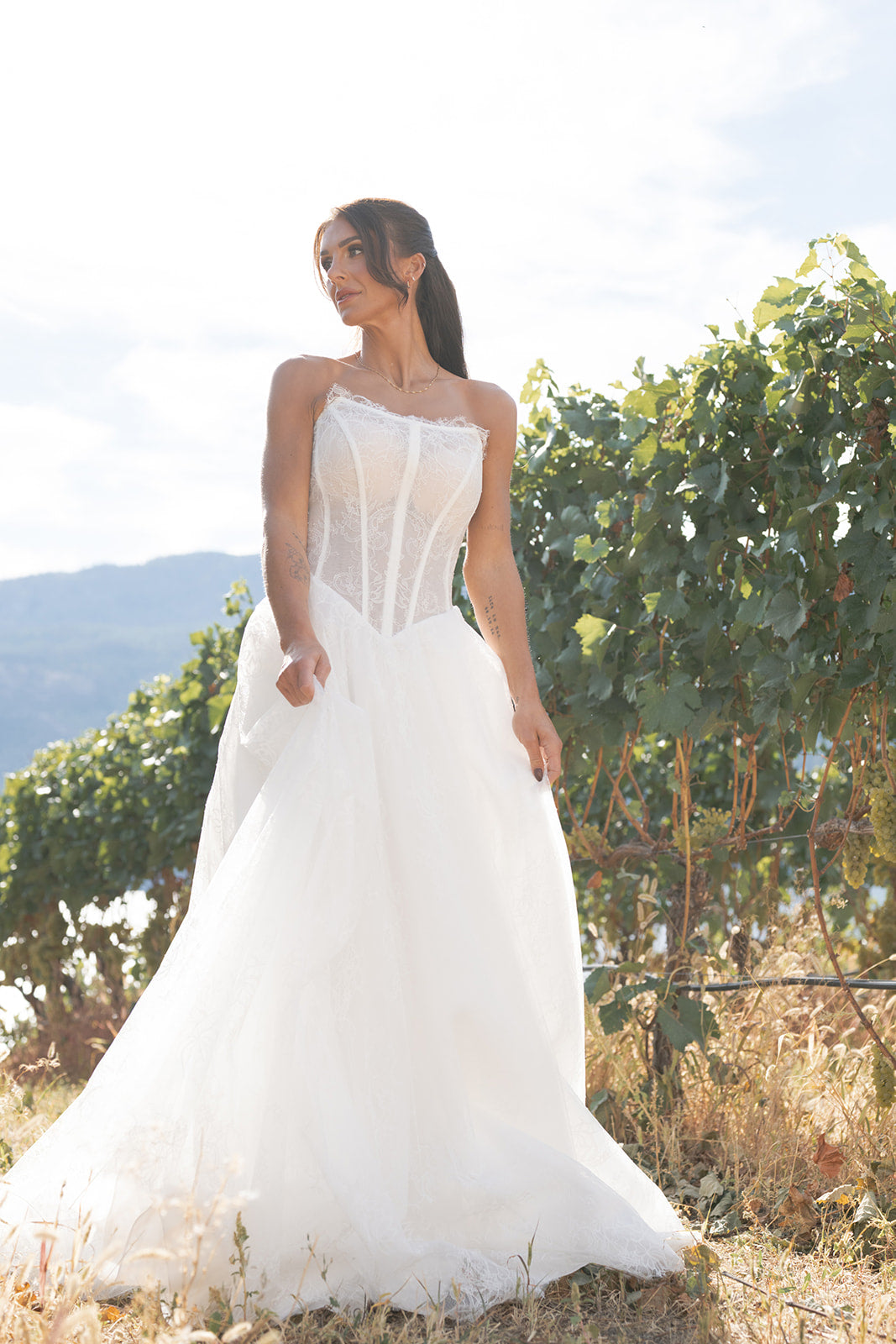 Woman in a white Amanti Contessa Strapless cat-eye neckline with exposed boning corset Basque waistline for a flattering, elongated shape wedding dress, standing in a vineyard with mountains in the background. Le Reve Boutique, St. Albert Alberta
