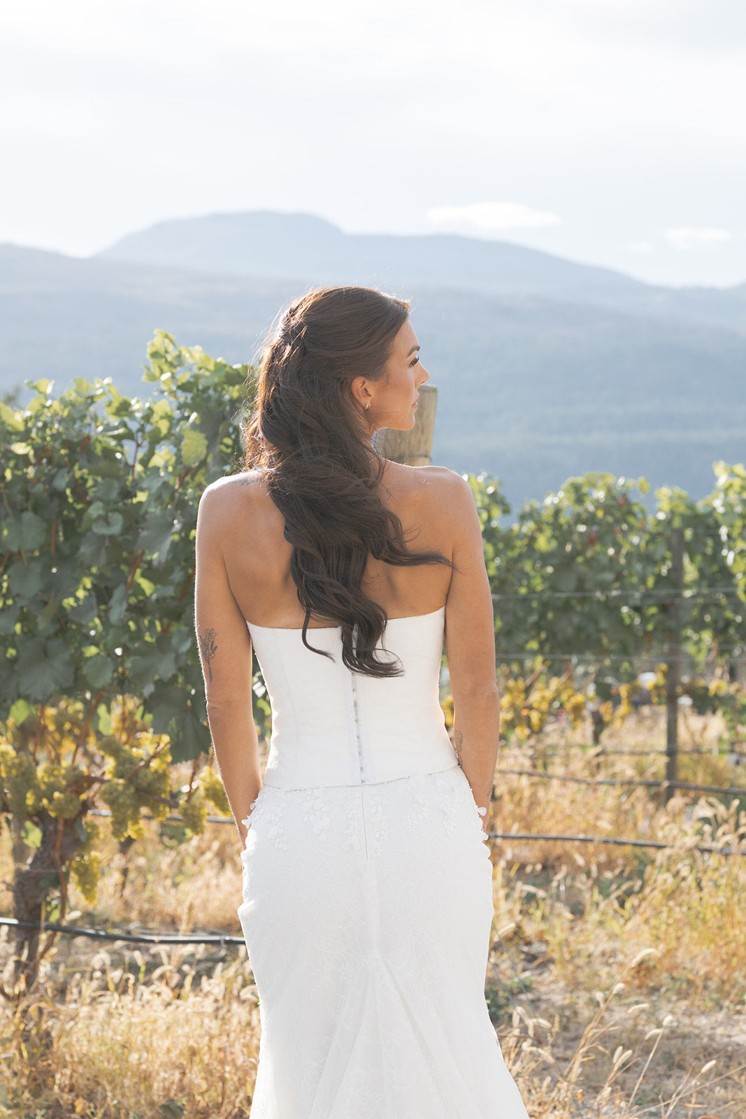 Woman in a white Satin corset with exposed boning, Round neckline with structured fit, Hook-and-eye back closure dress standing in a vineyard with mountains in the background.  Amanti at Le Reve Boutique. St. Albert, Alberta