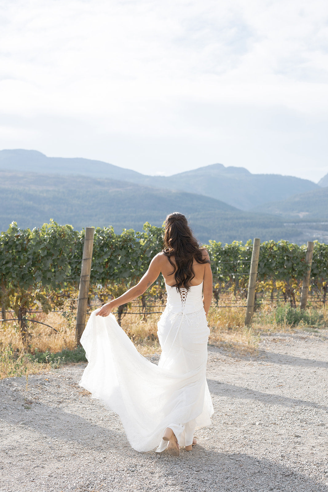 Woman in an Amanti Fiorina Set white Two-piece bridal set: satin bodice + Chantilly lace skirt, Lace-up corset back for a secure, adjustable fit. Hand-sewn floral appliqué details on the bodice, with soft floral Chantilly lace skirt with romantic texture wedding dress walking on a dirt path with mountains in the background. Le Reve Boutique