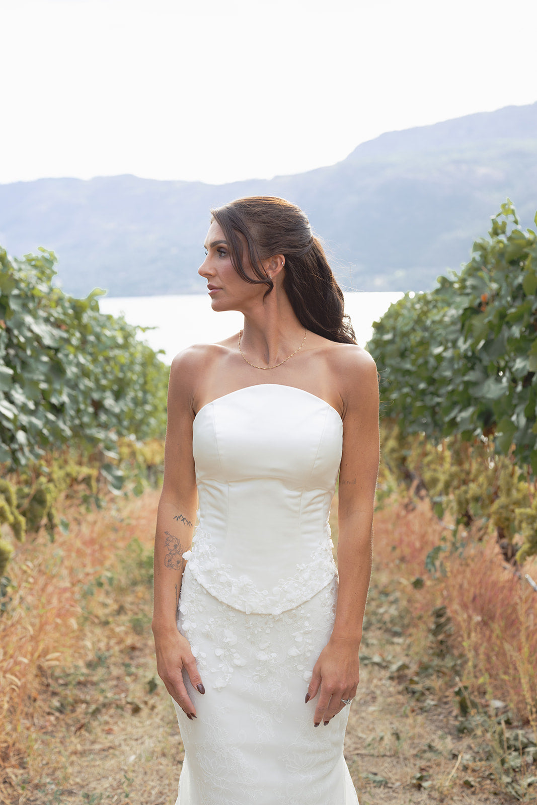 Woman in an Amanti Fiorina Set white strapless Hand-sewn floral appliqué details on the bodice wedding dress standing in a natural setting with mountains and greenery. Le Reve Boutique.  St. Albert, Alberta
