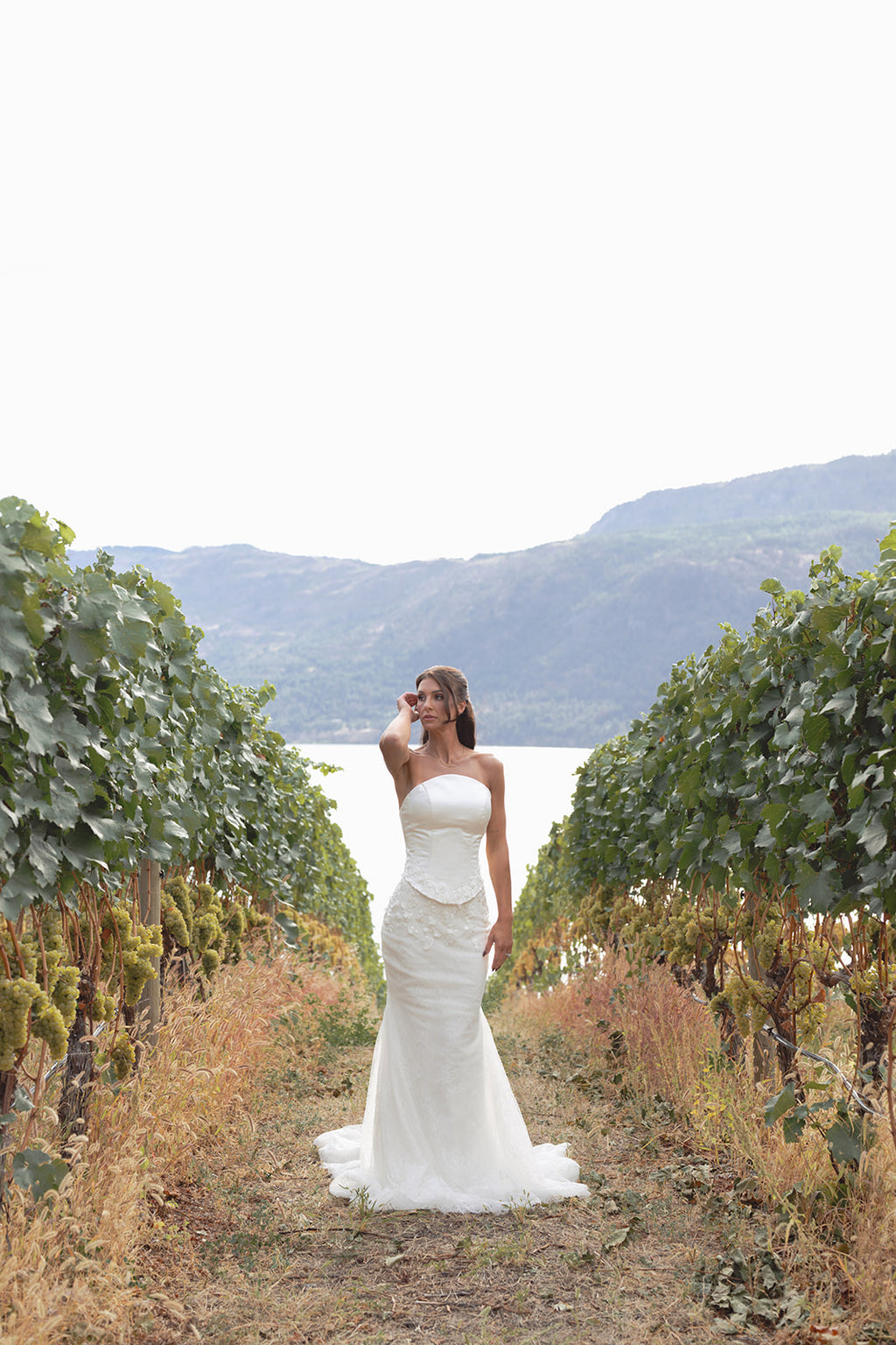 Woman in an Amanti Fiorina Set white Two-piece bridal set: satin bodice + Chantilly lace skirt wedding dress standing in a vineyard with mountains in the background. Le Reve Boutique, St. Albert, Alberta.