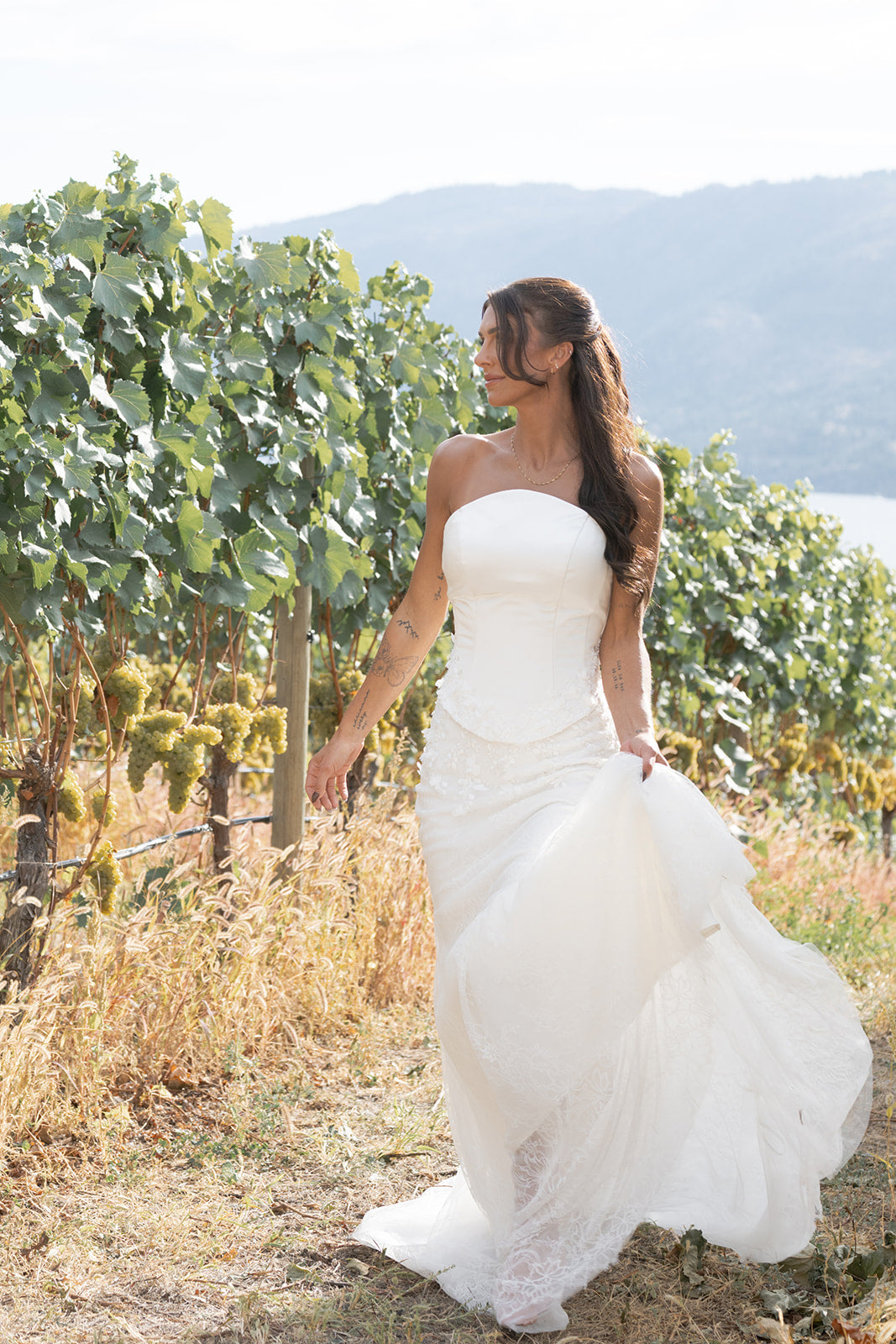 Woman in an Amanti Fiorina Set white Two-piece bridal set: satin bodice + Chantilly lace skirt, Lace-up corset back for a secure, adjustable fit, Hand-sewn floral appliqué details on the bodice, Soft floral Chantilly lace skirt with romantic texture wedding dress standing in a vineyard with mountains in the background.  Le Reve Boutique