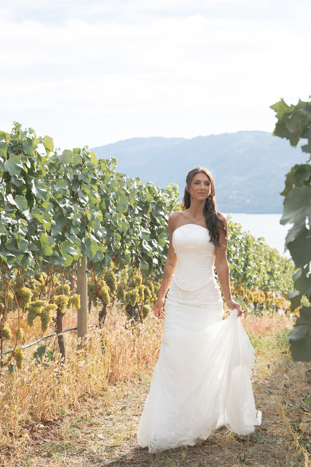 Woman in an Amanti Fiorina Set white wedding dress standing in a vineyard with mountains in the background.  This two-piece bridal gown offers a modern twist on timeless elegance. The structured satin bodice features a lace-up corset back for a customizable fit, creating a sleek and defined silhouette. Hand-sewn floral appliqué details add a touch of romance and texture, elevating the simplicity of the satin top.  Le Reve Boutique