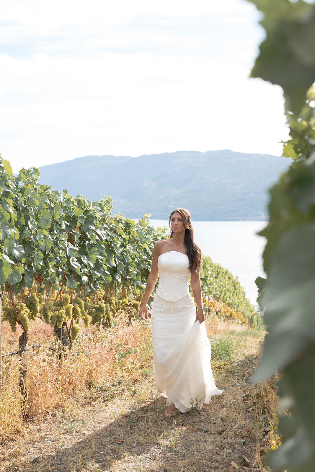 Woman in an Amanti Fiorina Set white wedding dress walking through a vineyard with mountains in the background. Skirt crafted in delicate floral Chantilly lace, flowing gracefully to the floor with soft movement and dimension. Together, the pieces create a look that feels fresh, stylish, and unforgettable — perfect for the bride who loves classic bridal style with a fashion-forward edge.  Le Reve Boutique Edmonton, Alberta