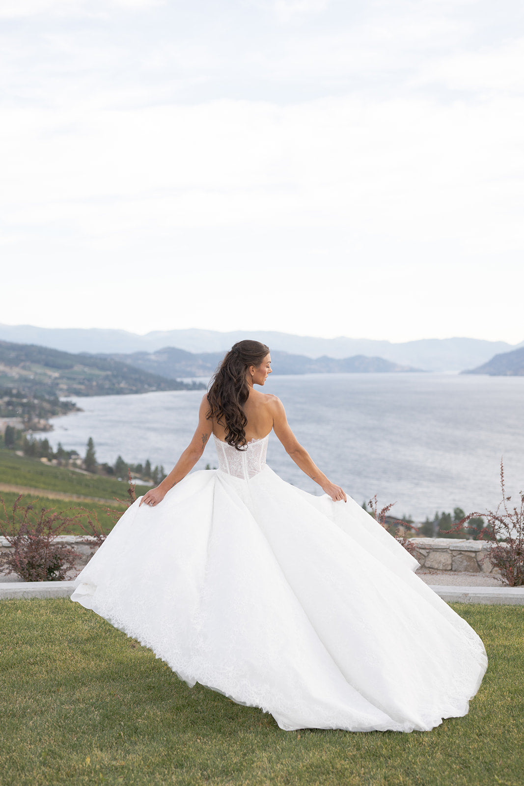 Woman in Amanti Fortuna A-line wedding dress, standing on grass with a scenic lake and mountains in the background.  Crafted in delicate French Chantilly lace. Straight across neckline elevated with a subtle keyhole detail, structured bodice, basque waistline . Le Reve Boutique, Edmonton, Alberta