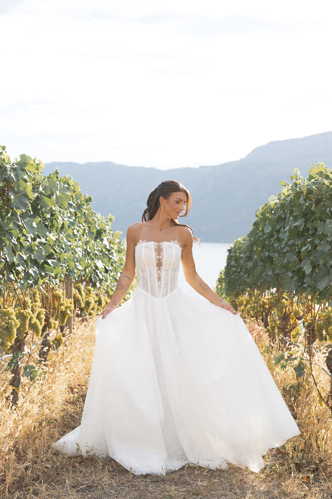 Woman in a Amanti Fortuna white Straight across neckline with keyhole detail Basque waistline for a flattering, elongated shape wedding dress standing in a vineyard with mountains in the background.  Le Reve Boutique St. Albert, Alberta