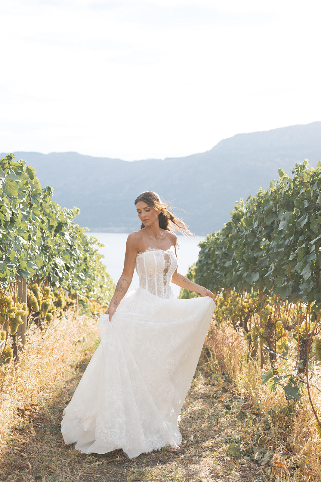 Woman in a white Straight across neckline with keyhole detail, Basque waistline for a flattering, elongated shape, A-line silhouette in Chantilly French lace,  for the modern bride, clean lines and comfort dress standing in a vineyard with mountains in the background. Amanti Fortuna, Le Reve Boutique Edmonton