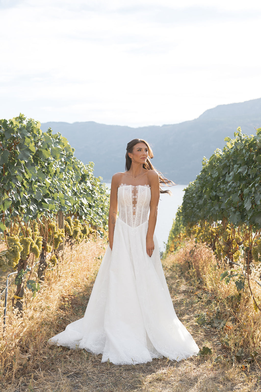 Woman in a white Straight across neckline with keyhole detail, Basque waistline for a flattering, elongated shape, A-line silhouette in Chantilly French lace wedding dress standing in a vineyard with mountains in the background.Amanti Fortuna Le Reve Boutique, Edmonton, Alberta