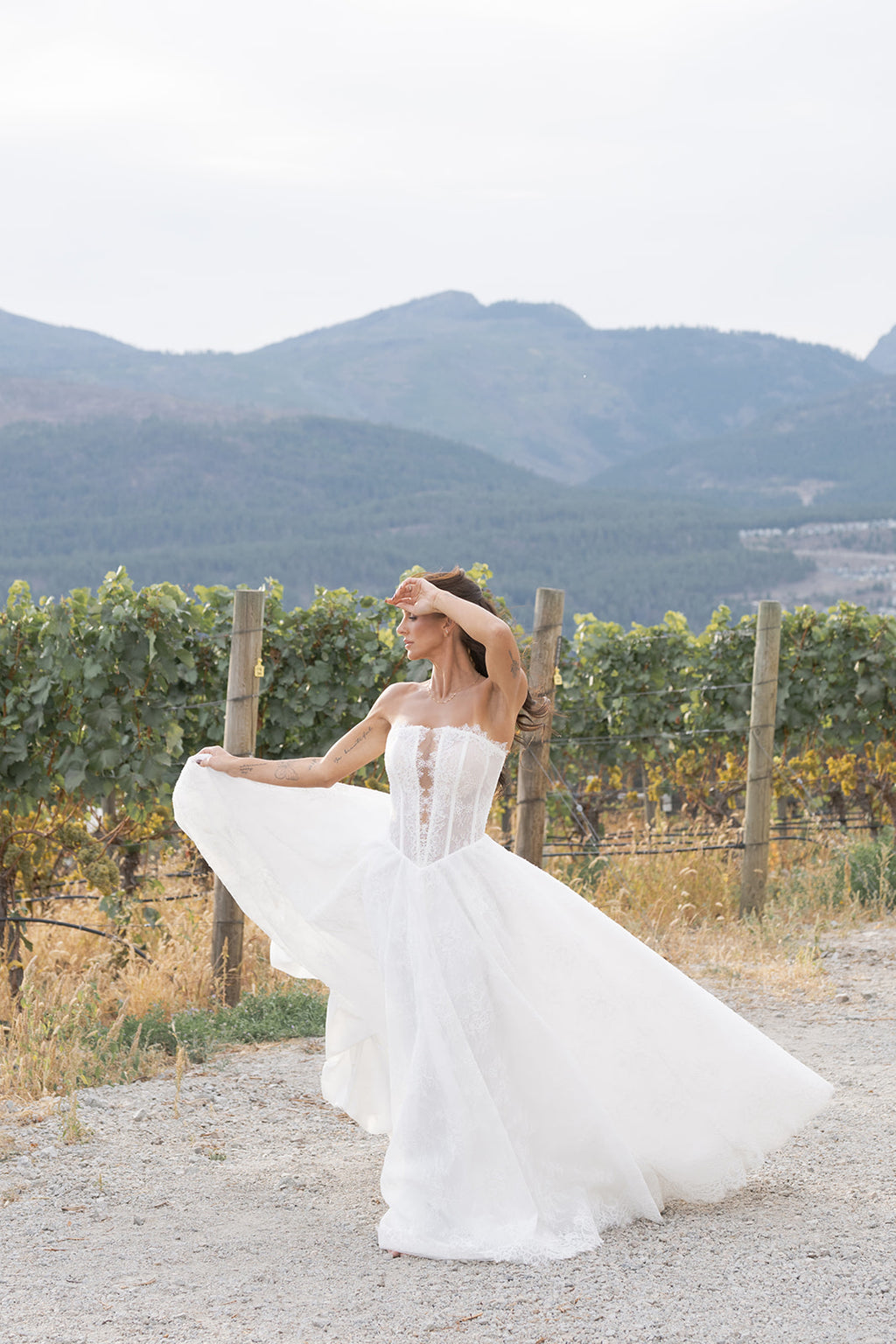 Woman in a white Straight across neckline with keyhole detail, Basque waistline for a flattering, elongated shape, A-line silhouette in Chantilly French lace, Zipper back closure for clean lines and comfort dress standing in a vineyard with mountains in the background. Amanti Fortuna, Le Reve Boutique Edmonton