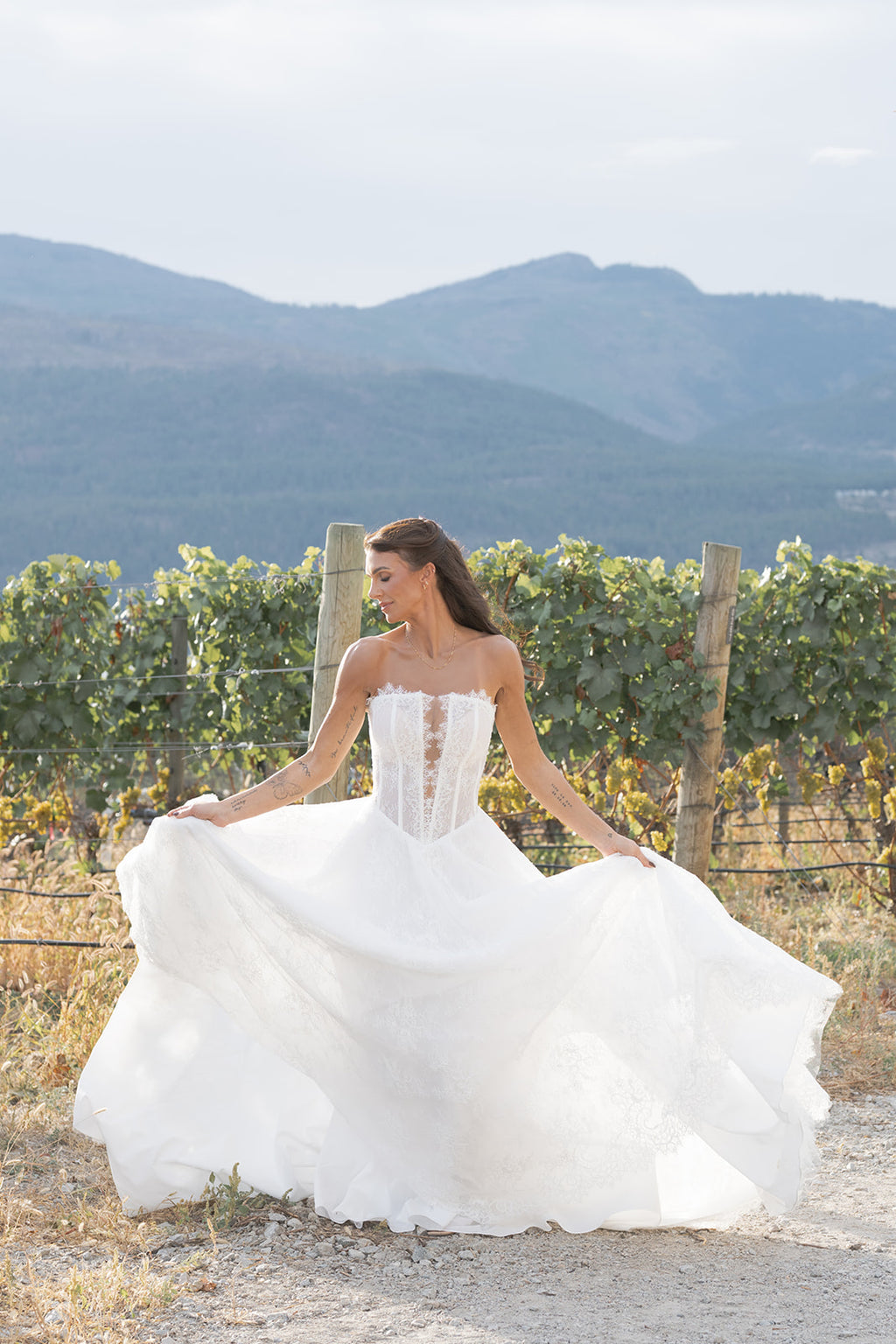 Woman sitting in Amanti Fortuna white wedding dress with Straight across neckline with keyhole detail, Basque waistline for a flattering, elongated shape, A-line silhouette in Chantilly French lace. vineyard with mountains in the background. Le Reve Boutique St. Albert, Alberta
