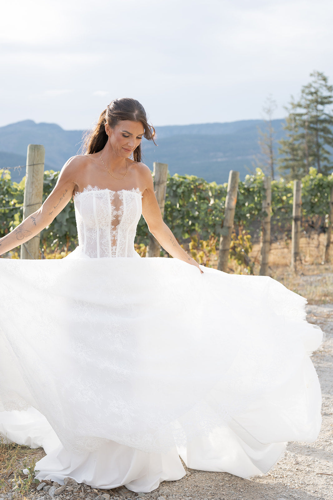 Woman in an Amanti Fortuna white wedding dress standing in a scenic outdoor setting with mountains and trees.  The zipper back closure keeps the design sleek and secure, while the lightweight lace layers move beautifully with every step. This gown is a stunning choice for brides who want elegance, comfort, and unforgettable detail in their wedding day look. Le Reve Boutique Edmonton, Alberta