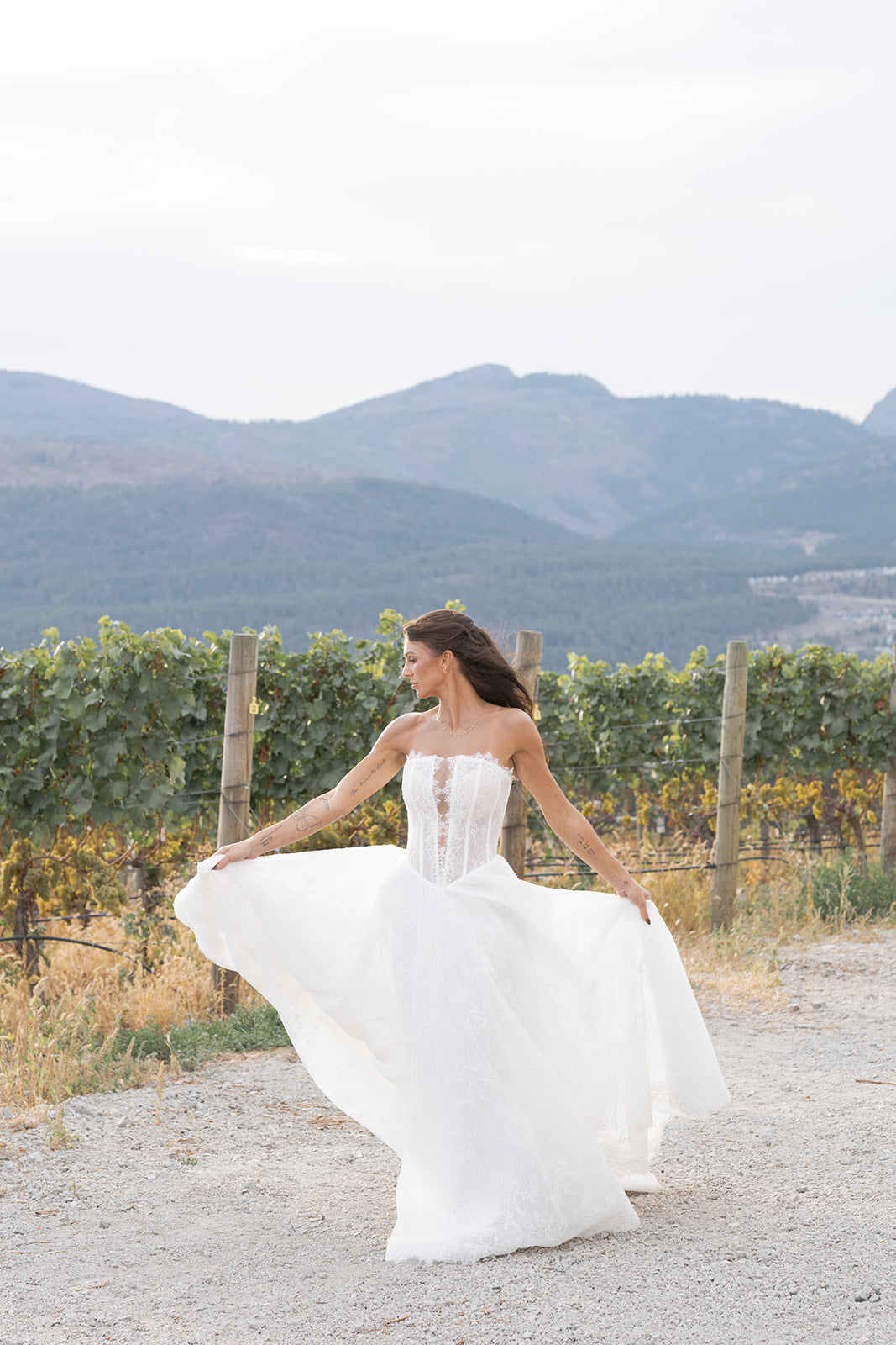 Woman in a full skirt Amanti Fortuna white wedding dress with Straight across neckline with keyhole detail, Basque waistline for a flattering, elongated shape, A-line silhouette in Chantilly French lace. vineyard with mountains in the background. Le Reve Boutique St. Albert, Alberta