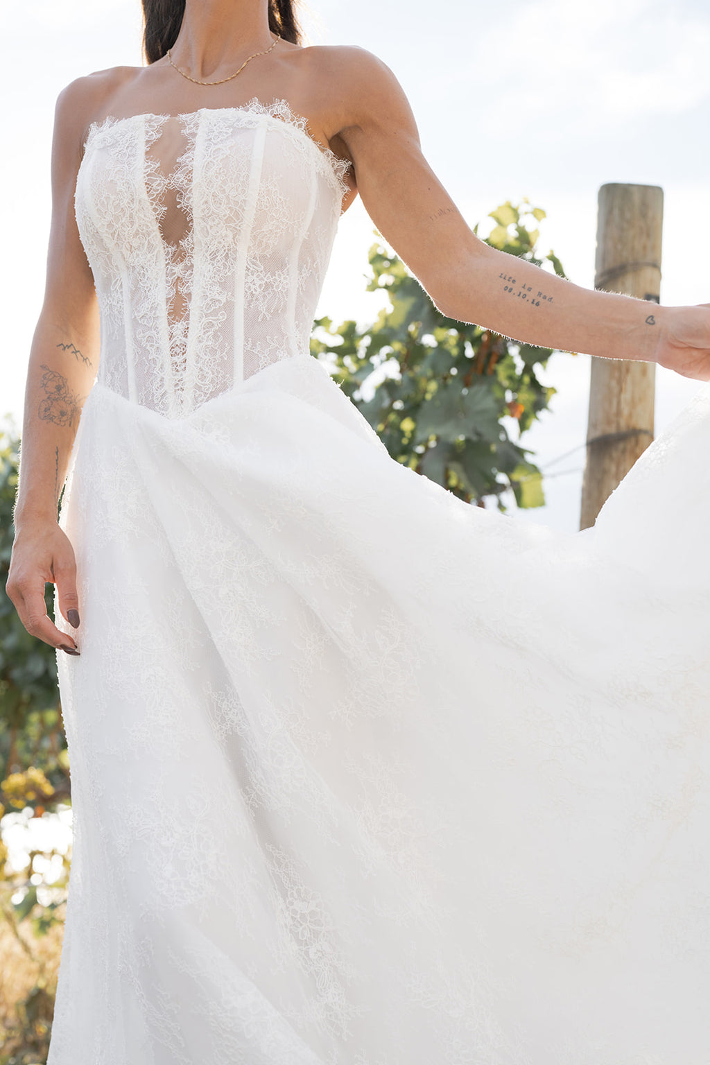 Woman in a  Amanti Fortuna white wedding dress with Straight across neckline with keyhole detail, Basque waistline for a flattering, elongated shape, A-line silhouette in Chantilly French lace, Zipper back closure for clean lines and comfort dress standing in a vineyard. Le Reve Boutique Edmonton White strapless lace wedding dress with a blurred natural background